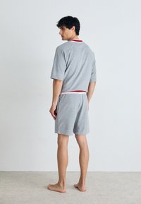 Grau, kurzärmliges Oberteil mit rotem Saum und passender grauer Shorts, aus weichem Stoff. Das Model steht barfuß, von hinten gewandt, mit sichtbarer Handytasche.