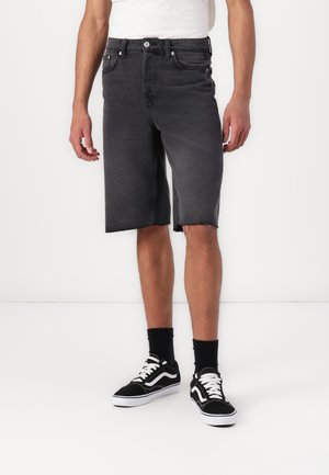 Grå denimshorts med frynset kant, fem lommer, frontlukking med knapp, brukt med svarte sneakers og svarte ankelstrømper mot en nøytral bakgrunn.