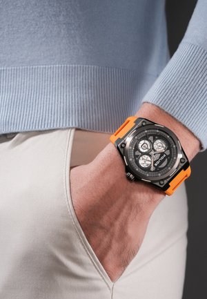 Vyras dėvi juodą ir oranžinį chronografinį rankinį laikrodį su keliais ciferblatais, ranka į kišenę įkišta į smėlio spalvos kelnes, matoma šviesiai mėlyno megztinio rankovė.