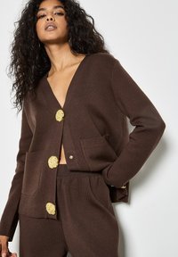 Cardigan marrone lavorato a maglia con profondo scollo a V, bottoni dorati, due tasche frontali e maniche lunghe. Abbinato a pantaloni marroni coordinati.