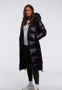 Lange zwarte pufferjas met capuchon, doorgestikte ontwerp, glanzende afwerking, ritssluiting aan de voorkant, zijzakken en een losse pasvorm, gedragen over een witte top en leggings.