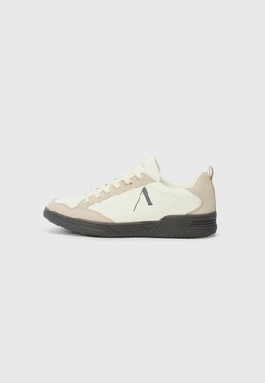 Niski sneaker z beżową cholewką ze skóry zamszowej i białej tkaniny, czarną gumową podeszwą, sznurowaniem z przodu oraz ciemnoszarym ukośnym logo na bocznej części.