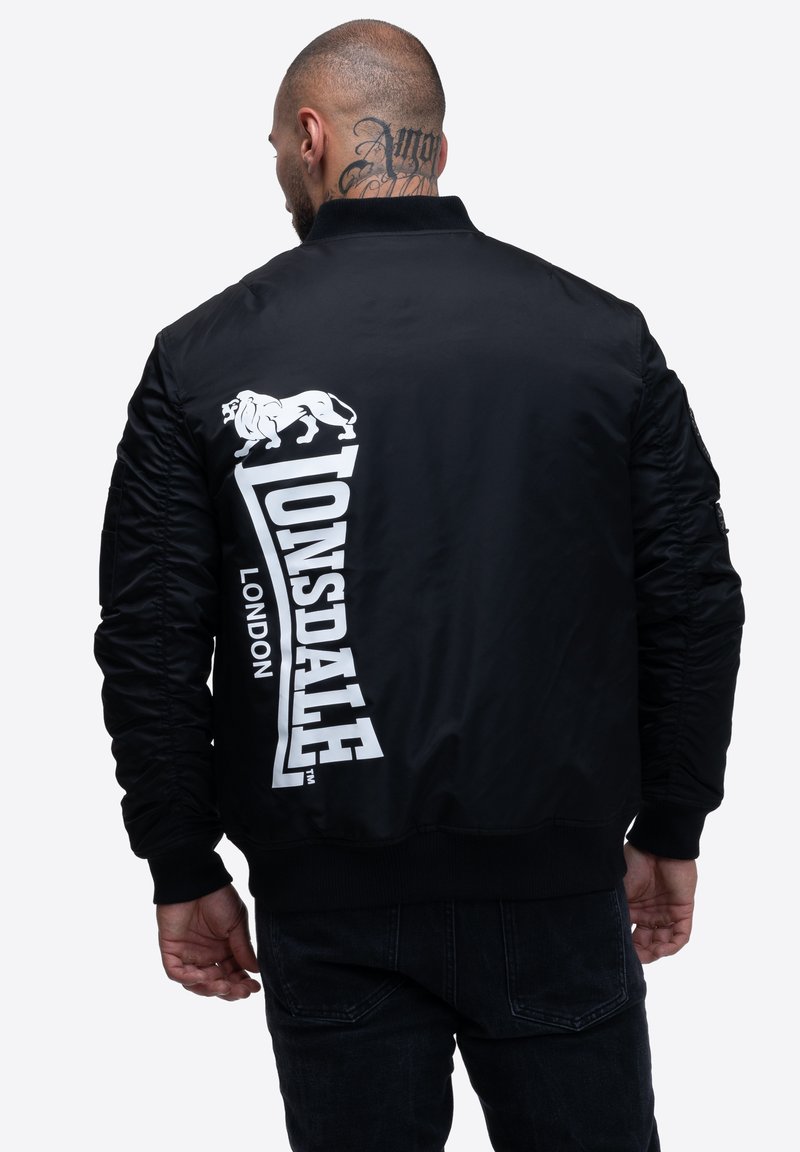 Chaqueta bomber negra con puños acanalados, que presenta un gran logo en blanco de "Lonsdale London" y un gráfico de león en la parte posterior. Textura de tela suave.