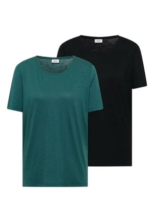 Femme souriante aux longs cheveux bruns portant un t-shirt à manches courtes couleur sarcelle et une ceinture beige clair avec un jean bleu sur un fond clair.