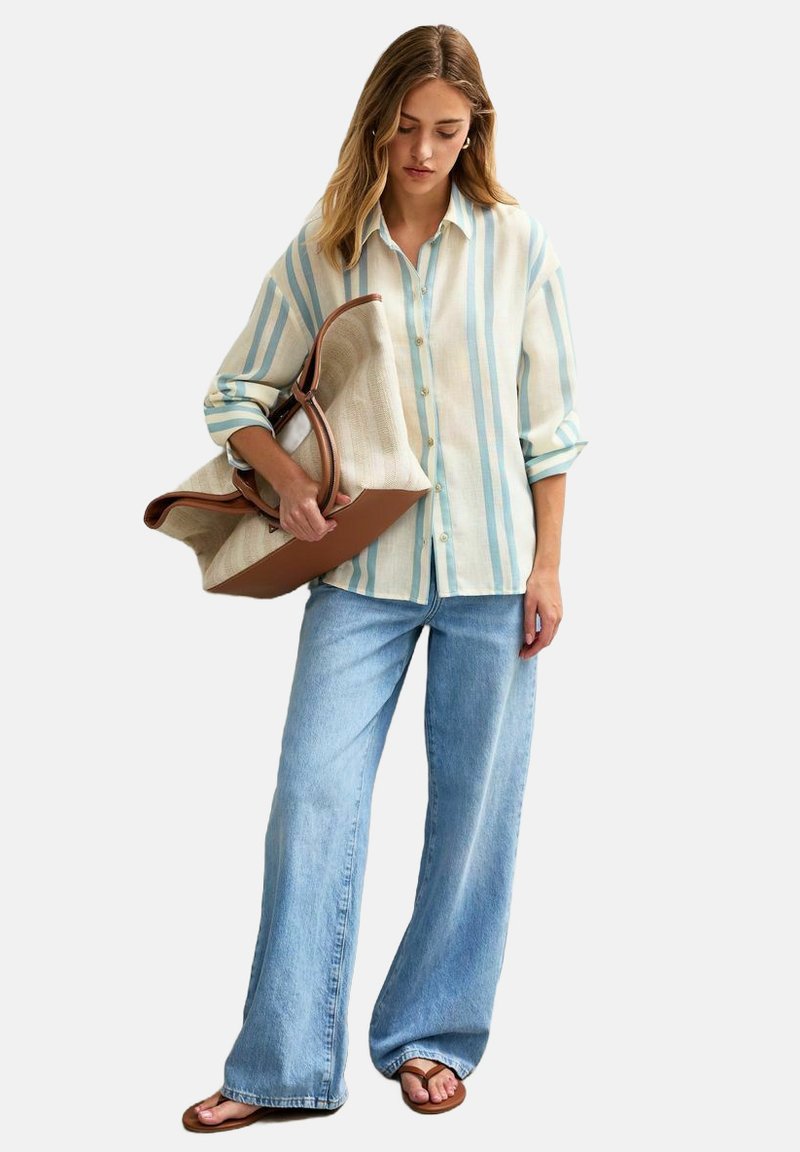 Camicia oversized a righe chiare in morbido tessuto, righe blu e beige, con bottoni sul davanti. Jeans in denim a vestibilità ampia, sandali piatti, grande borsa marrone.