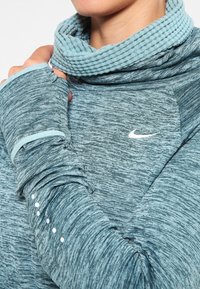 Türkisfarbenes Langarmshirt mit geripptem Rollkragen, strukturierter Stoff und Daumenlöchern. Mit Nike-Logo und weißen Akzentpunkten an den Bündchen.