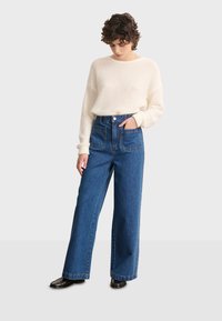 Bijela rebrasta pletena majica u kombinaciji s visokim širokim plavim trapericama koje imaju dva prednja džepa i kopčanje na gumb. Crne cipele.