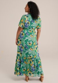 Robe maxi florale verte à manches courtes, ornée de motifs violets et jaunes, avec un ourlet à volants et une ceinture en tissu à la taille.