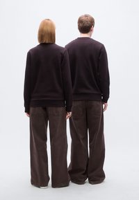 Δύο άτομα που φορούν σκούρα καφέ φούτερ και καφέ παντελόνια με φαρδιά πόδια, από πίσω, πάνω σε ανοιχτό γκρι φόντο.