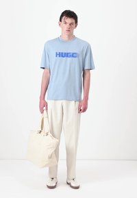 Light blue cotton t-shirt with "HUGO" logo in darker blue, paired with cream tapered trousers and a large cream fabric tote bag.
