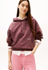 Sweat à capuche bordeaux avec poche avant, capuche à cordon de serrage et poignets côtelés, porté sur une chemise blanche. Associé à un pantalon en velours côtelé rose.