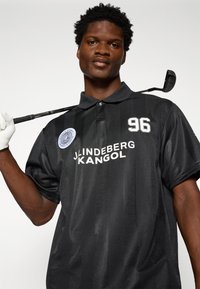 Sort poloshirt med korte ærmer og en glat tekstur. Har et stribet design, hvidt logo og nummer "96" på fronten. Golfkølle holdt i hånden.