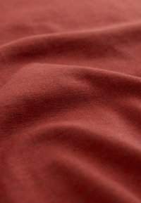 Rozsda színű pamutszövet sima textúrával és finom rugalmassággal, lágy, hullámzó redőkkel és enyhe árnyalatkülönbségekkel.