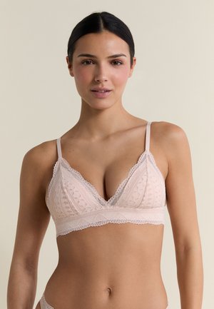 Woman with dark hair wears a light pink lace bralette with thin straps against a plain beige background.