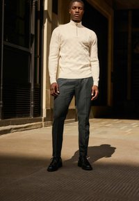 Jersey de punto en color crema con cremallera lateral, combinado con pantalones oscuros de corte ajustado en gris y zapatos de cuero negros, de pie sobre una superficie de piedra.