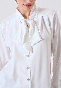 Witte blouse met een gestrikte hals, zachte stof, lange mouwen en goudkleurige knopen aan de voorkant. Gladde textuur en een schone uitstraling.