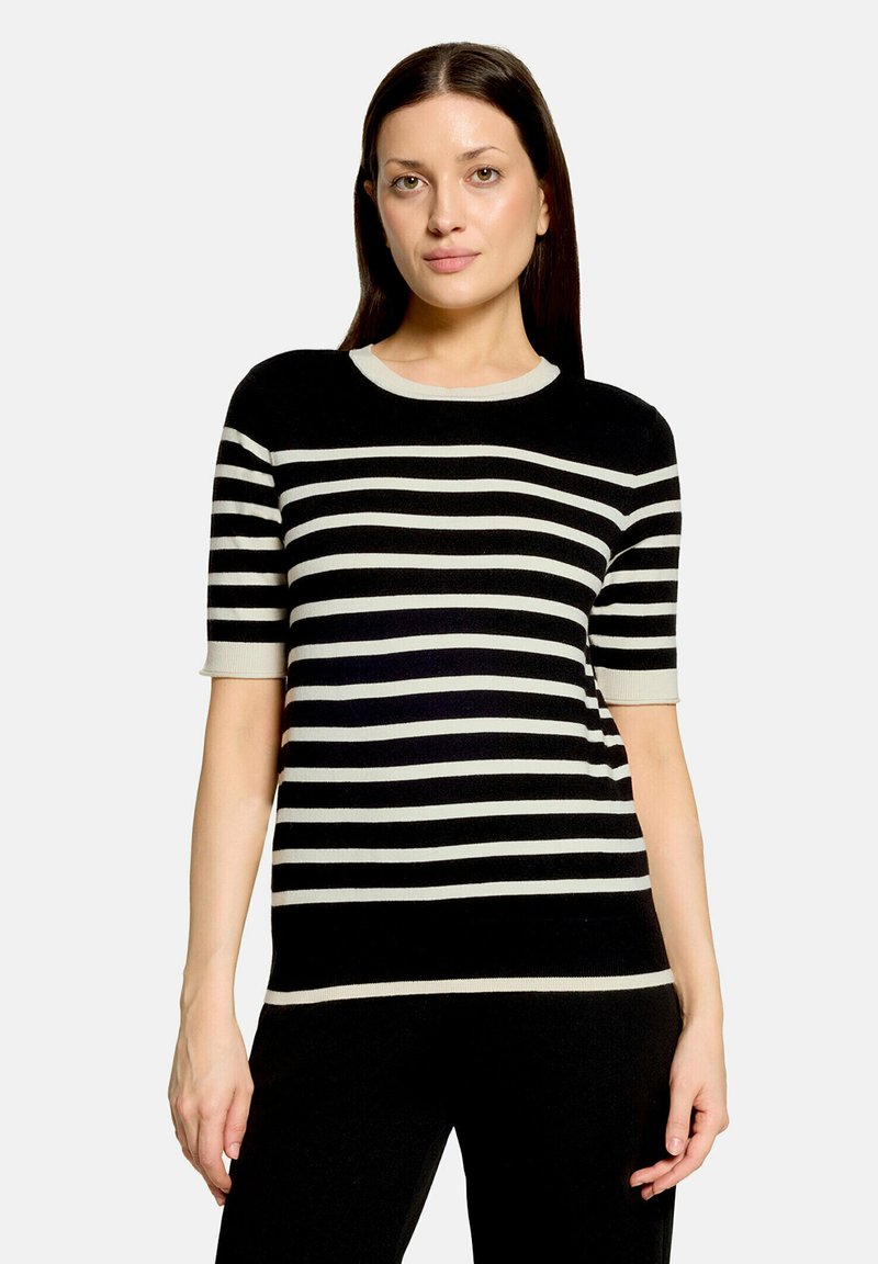 Femme aux cheveux lisses et foncés portant un pull noir à manches courtes avec des rayures blanches horizontales sur un fond clair.