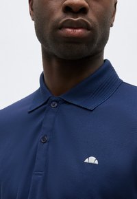 Gros plan d'un homme portant un polo bleu marine texturé avec une patte de boutonnage à trois boutons et un petit logo blanc en forme de demi-cercle sur la poitrine.