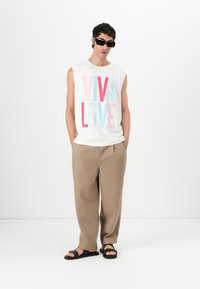 Hvit ærmeløs skjorte med "VIVA LOVE" grafikk i rosa og blå, beige løstsittende bukser og svarte sandaler. Enkelt, uformelt design.
