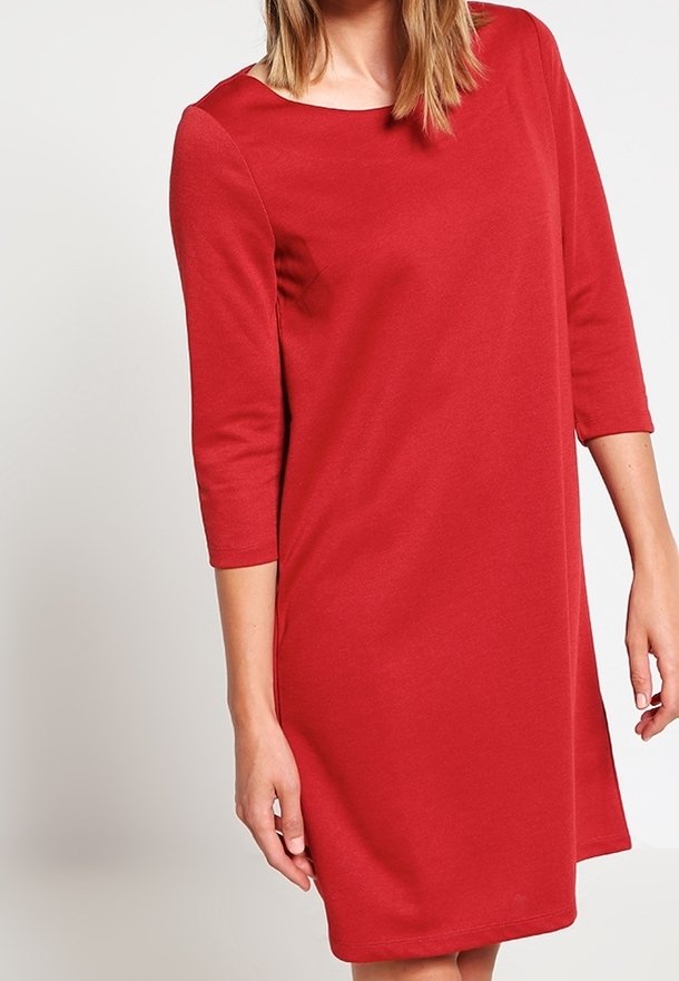 Femme portant une robe rouge mi-longue avec des manches trois-quarts et un col rond, debout devant un fond clair uni.