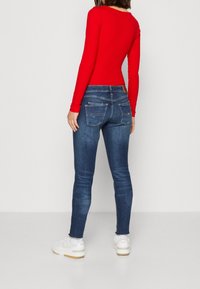 Top rojo de manga larga combinado con unos jeans slim-fit de color azul oscuro. Los jeans tienen bolsillos traseros y un parche de cuero marrón en la cinturilla. Zapatillas blancas.