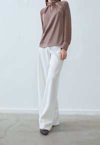 Seidige braune Bluse mit einem Schlitzdetail, kombiniert mit hoch taillierten, weit geschnittenen weißen Hosen und spitzen schwarzen Absätzen auf einem Betonboden.