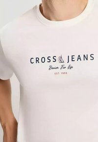 Biała koszulka polo z krótkim rękawem z logo "CROSS JEANS", wzorem żaglówki oraz napisem "Denim For Life EST. 1990" w granatowym i czerwonym kolorze.