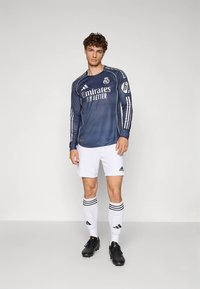 Marineblauw voetbalshirt met lange mouwen, witte strepen en branding, gecombineerd met witte korte broek en zwarte voetbalschoenen, met drie strepen op de sokken.