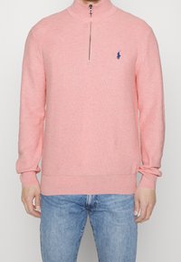 Rosa stickad tröja med hög krage, försedd med en halv dragkedja och en liten blå logga. Texturen är mjuk och ärmarna sitter tight.