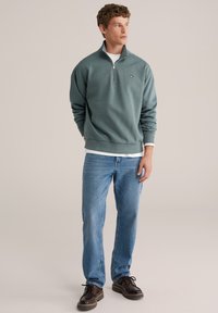 Zelen pulover s četrtinskimi zadrgami, rebrastimi manšetami in robom, oblečen čez belo srajco, kombiniran s modrimi kavbojkami in rjavimi čipkastimi čevlji.