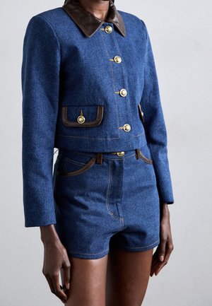 Person trägt eine blaue Wolljacke mit braunem Kragen und goldenen Knöpfen, kombiniert mit passenden blauen hoch taillierten Shorts mit braunem Rand.