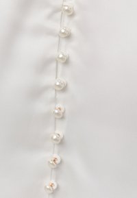 Tissu blanc avec une rangée verticale de petits boutons en nacre cousus avec du fil blanc et des pieds dorés.