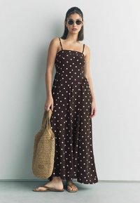 Brązowa sukienka maxi w kropki z cienkimi ramiączkami, plisowanym dołem i plecioną beżową torbą. Modelka nosi czarne okulary w okrągłej oprawie i sandały.