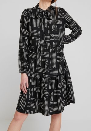 Schwarzes langärmliges Kleid mit weißen geometrischen Pfeilmustern, einer Schleifenbindung am Hals und einem gestuften Rock, getragen von einer Frau vor schlichtem Hintergrund.
