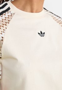 Krämfärgad sweatshirt med spetsdetaljer på axlarna. Har en liten svart logotyp på bröstet och randiga svarta detaljer på ärmarna.