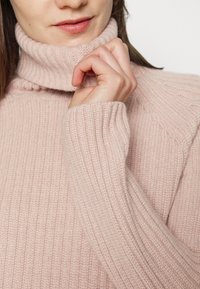 Femme portant un pull à col roulé côtelé rose clair, tenant délicatement le col d'une main près de ses lèvres.