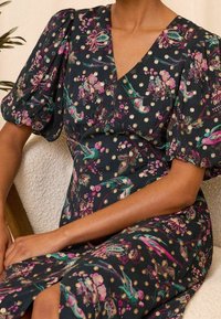 Blommönstrad klänning med mörk bakgrund, puffärm, v-ringning och en sidoklyvning. Har guldprickar och livfulla fåglar.