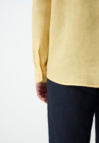 Camicia di lino gialla a maniche lunghe con polsi abbottonati, abbinata a pantaloni blu scuro testurizzati, che mostrano un primo piano dei dettagli del tessuto e dei bottoni.