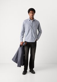 Gombos ing kék és fehér függőleges csíkokkal, hosszú ujjakkal, fehér gombokkal, sötét farmernadrággal párosítva, és egy szürke kabátot tartva.