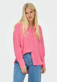 Camicia button-up rosa brillante con maniche lunghe, fit rilassato e texture morbida, abbinata a jeans blu a vita alta.