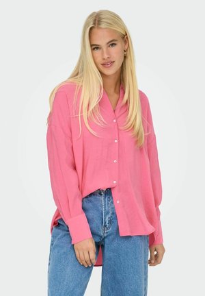 Camicia button-up rosa brillante con maniche lunghe, fit rilassato e texture morbida, abbinata a jeans blu a vita alta.