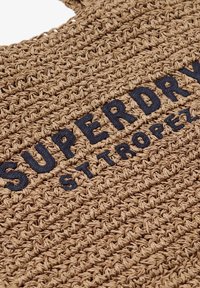 Bolso tote tejido en tono marrón claro con un diseño texturizado y el logo bordado en negro "SUPERDRY" y "ST TROPEZ" en la parte frontal.