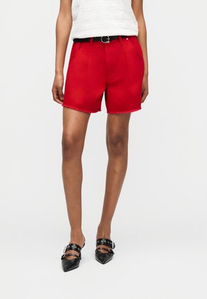 Μοντέλο που φοράει κόκκινα σορτς με μαύρη ζώνη, σε συνδυασμό με μαύρα παπούτσια μυτερά με λουριά και αγκράφες.