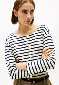 Maglietta bianca a maniche lunghe con righe orizzontali blu navy, dotata di scollo rotondo e sette bottoni dorati lungo la parte anteriore. Tessuto strutturato.