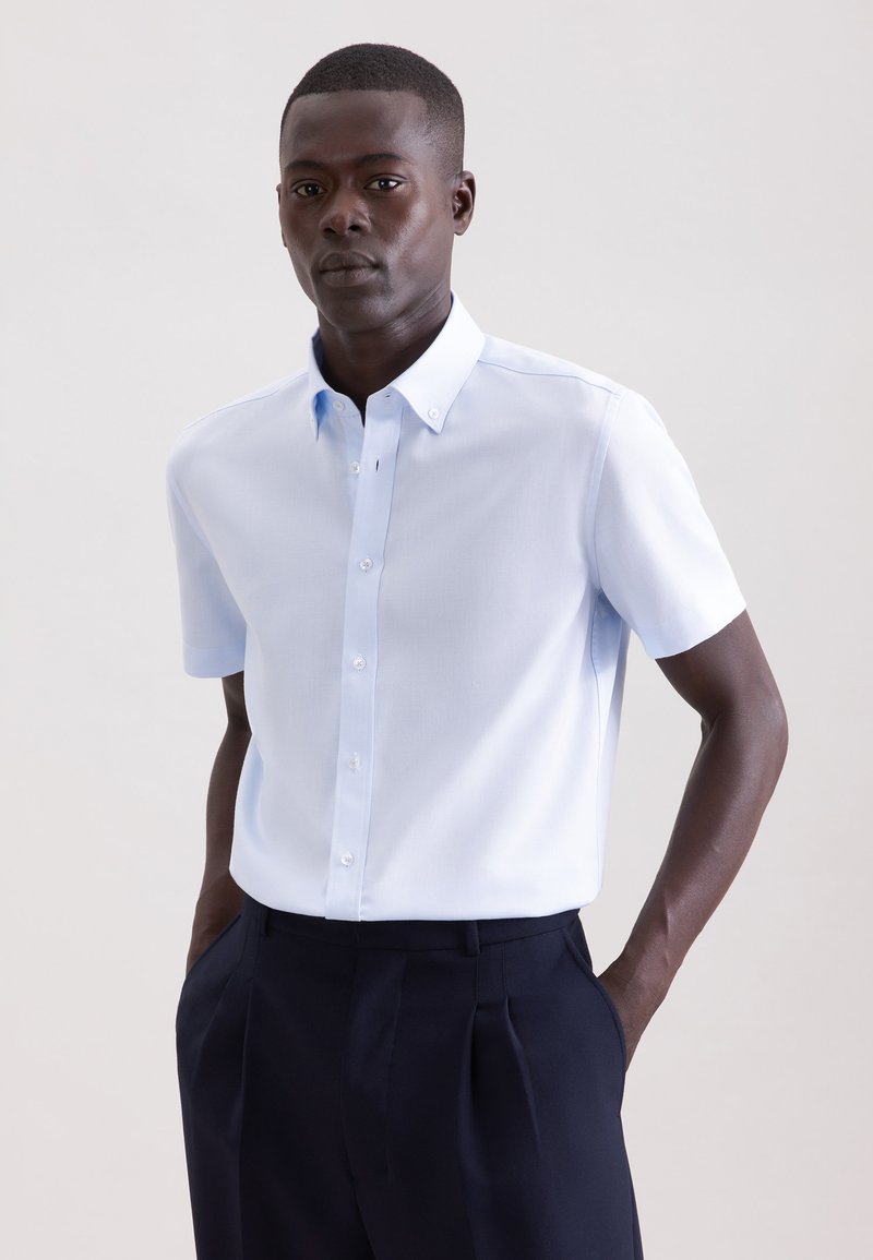 Homme portant une chemise à manches courtes bleu clair boutonnée, rentrée dans un pantalon plissé bleu marine foncé, debout les mains dans les poches.