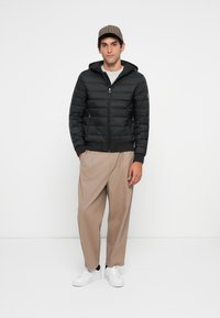 Young man wearing a black quilted jacket, beige wide-leg trousers, white sneakers, and a brown plaid cap, standing against a white background.