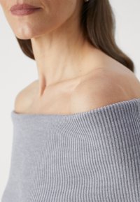 Maglione a coste grigio chiaro senza spalle, con un ampio scollo e una superficie liscia, con toni della pelle a contrasto visibili sulle spalle.