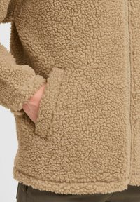 Veste en polaire beige avec un tissu texturé et duveteux. Dotée d'une fermeture éclair à l'avant et de poches latérales avec un ourlet lisse. Les poignets sont légèrement élastiqués.