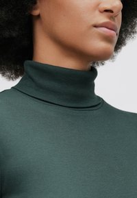 Nahaufnahme einer Person, die einen dunkelgrünen, gerippten Rollkragenpullover trägt, wobei Hals, Kieferlinie und Lippen vor einem schlichten Hintergrund zu sehen sind.