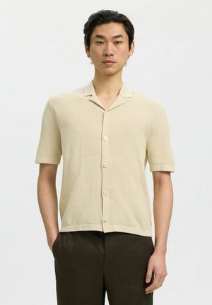Homme portant une chemise en maille beige à manches courtes boutonnée et un pantalon marron foncé, debout les mains dans les poches devant un fond uni.